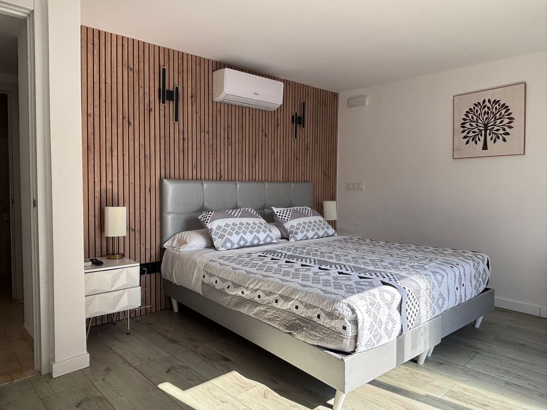 Dormitorio moderno con cama, pared decorativa de listones de madera, mesita de noche y lámina enmarcada de un árbol