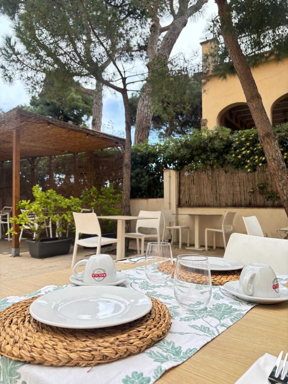 Mesa de café al aire libre con platos, tazas y copas de vino sobre un mantel individual tejido, bajo los árboles.