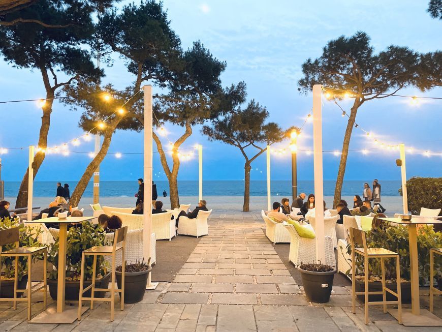 Cena en una terraza junto al mar al anochecer, con mesas iluminadas, palmeras y vistas al océano
