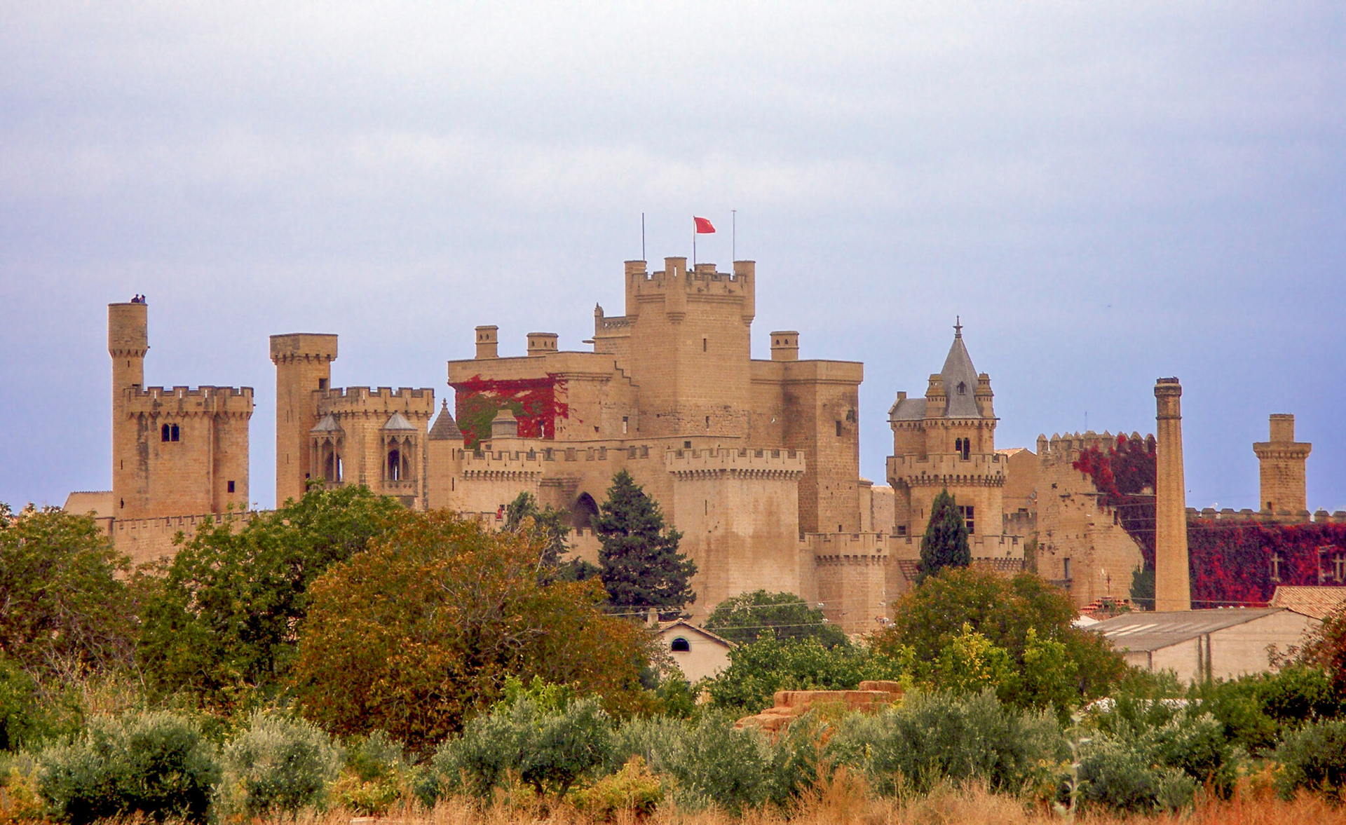 Castillo de arenisca color canela con torres y banderas rojas sobre árboles verdes bajo un cielo nublado.