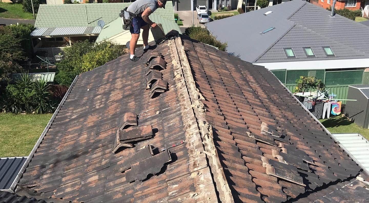 A worker repairs a damaged roof, replacing missing tiles along the ridge on a sunny day.
