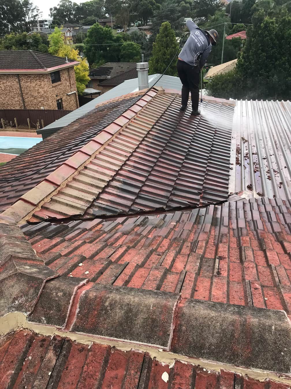 A person power washing a weathered terracotta-tiled roof on a residential building.