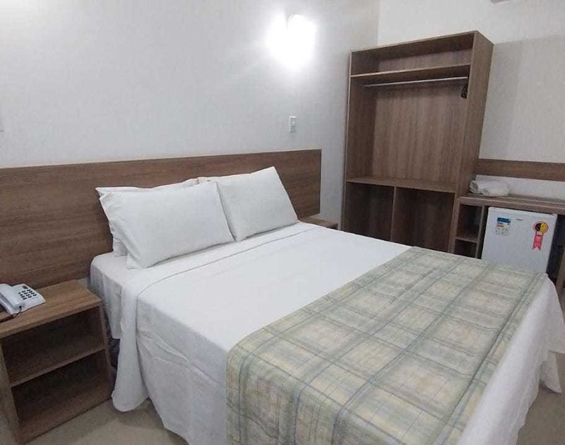 Quarto de hotel com cama de casal, armário, mesa de cabeceira e frigobar. Roupa de cama branca com manta xadrez.