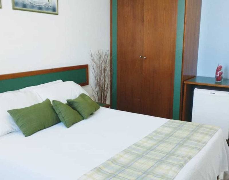 Quarto com roupa de cama branca, almofadas verdes, guarda-roupa de madeira e frigobar.