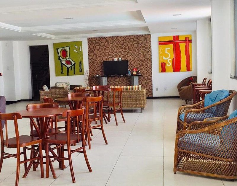 Cafeteria com mesas e cadeiras redondas de madeira, poltronas de vime, uma TV e obras de arte.