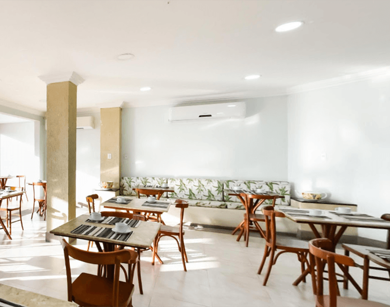 Área de jantar do restaurante com mesas e cadeiras de madeira, paredes brancas, um longo banco florido e ar condicionado.