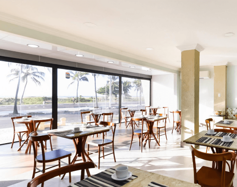 Interior de restaurante com mesas e cadeiras perto de uma grande janela com vista para uma praia com palmeiras.