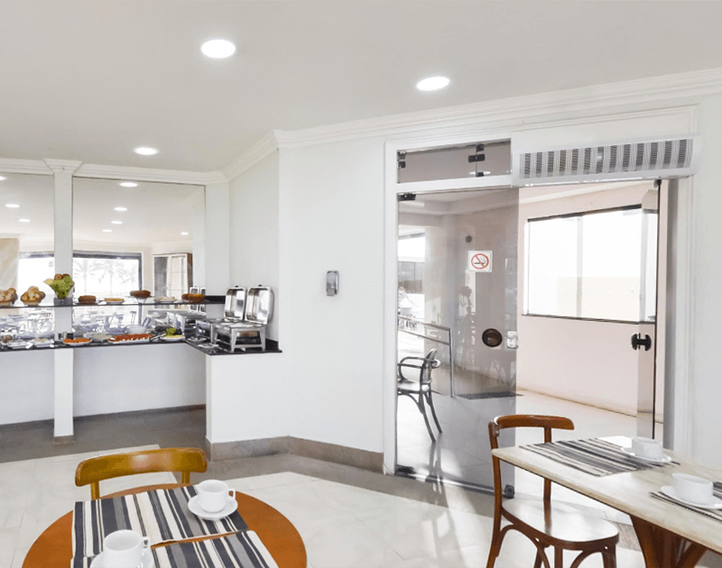 Área de café da manhã: buffet com opções de comida, mesas com cadeiras, porta de vidro, paredes brancas e iluminação clara.