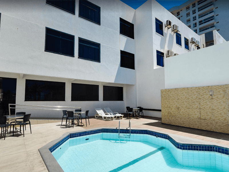 Exterior de hotel com piscina, mesas e cadeiras. Prédio branco com janelas escuras sob um céu azul.