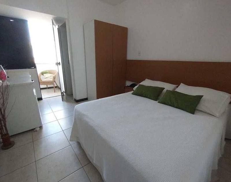 Quarto com cama, guarda-roupa e acesso à varanda. Almofadas verdes e roupa de cama branca.