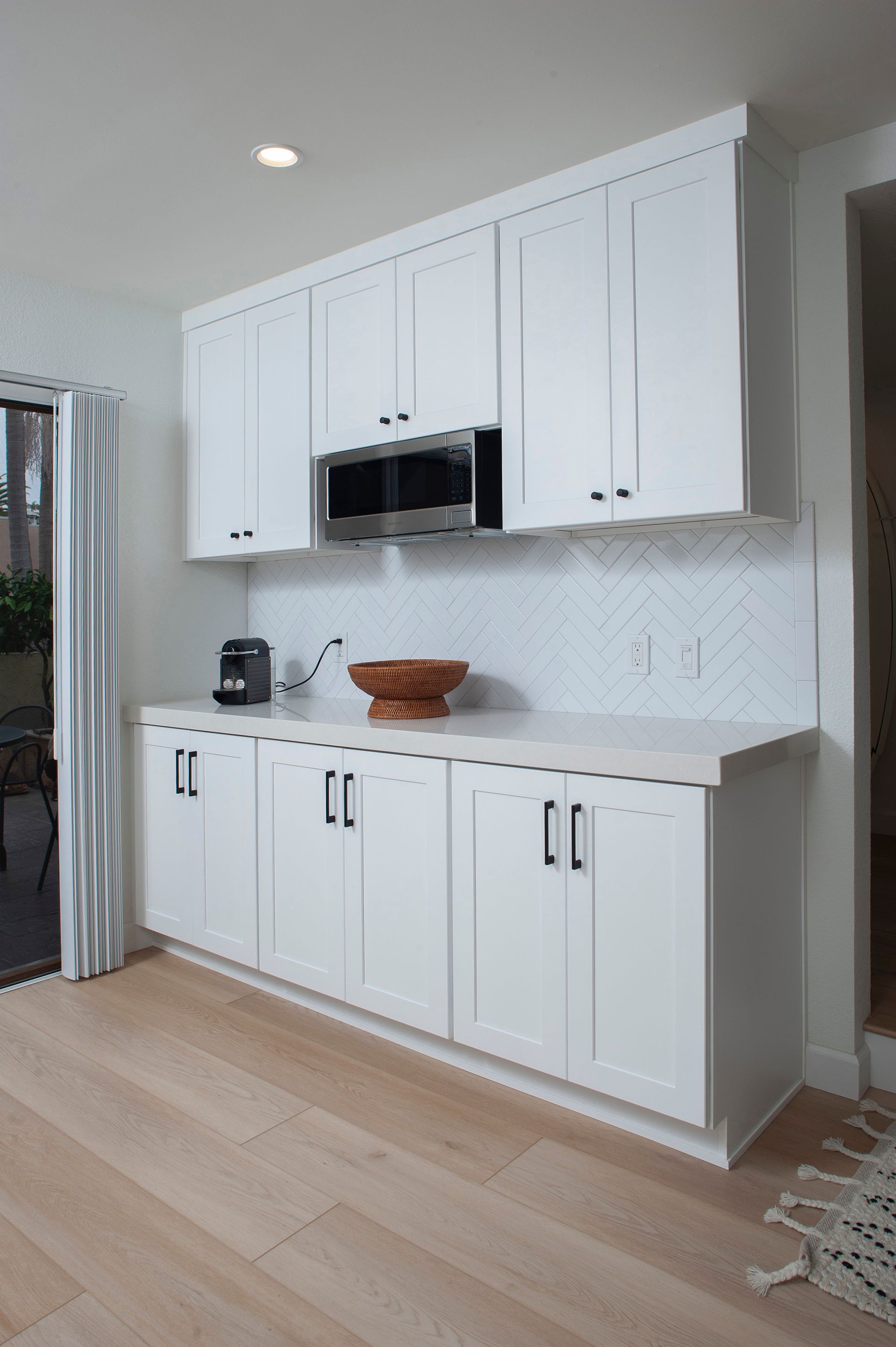 White kitchenette with cabinets, microwave, and sink. Wooden floor and herringbone backsplash.