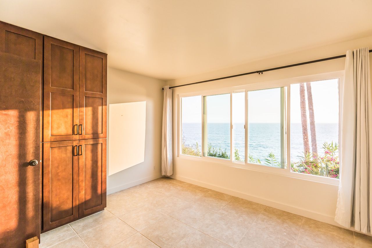 Empty room with ocean view, beige walls, wooden cabinets, large window, and white curtains.