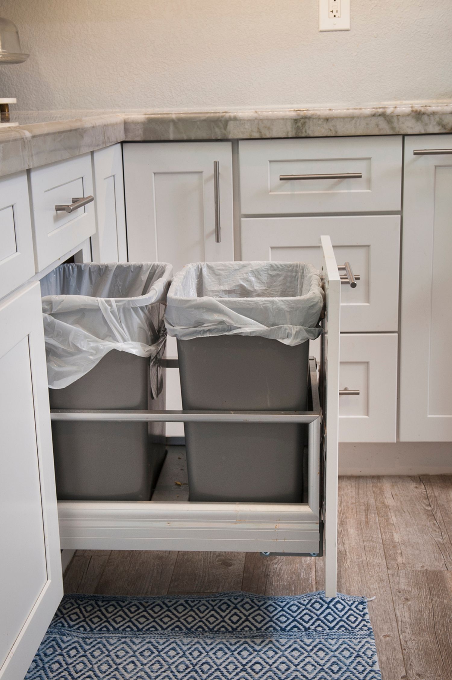 A pull-out kitchen cabinet with two trash bins. White cabinets, grey bins with liners, a blue rug.