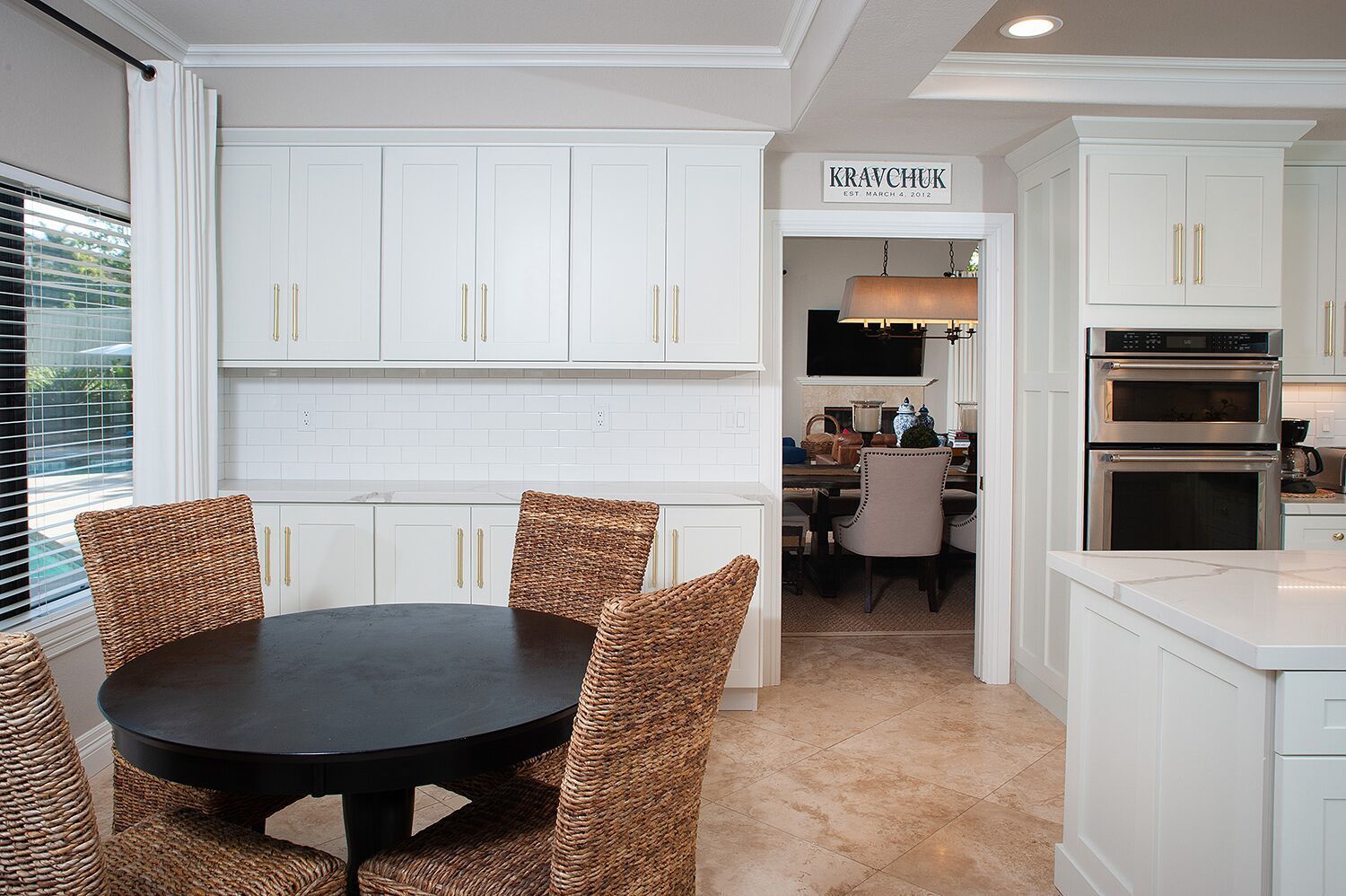 A kitchen with white cabinets, dark round table, woven chairs, and a view into a dining room.