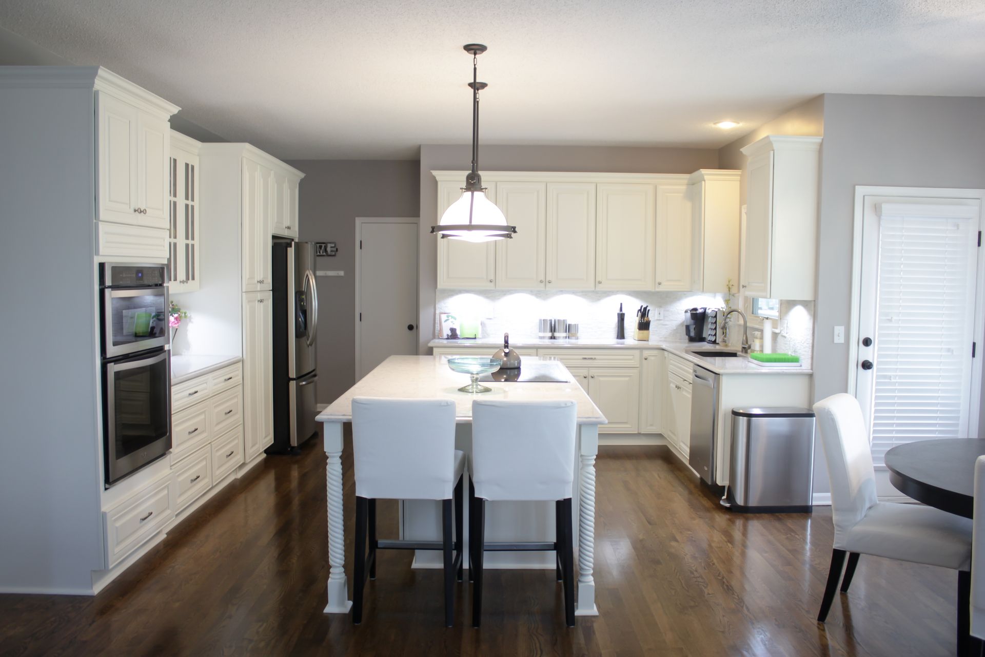Bright kitchen with white cabinets, island, and chairs; a dark hardwood floor; and stainless steel appliances.