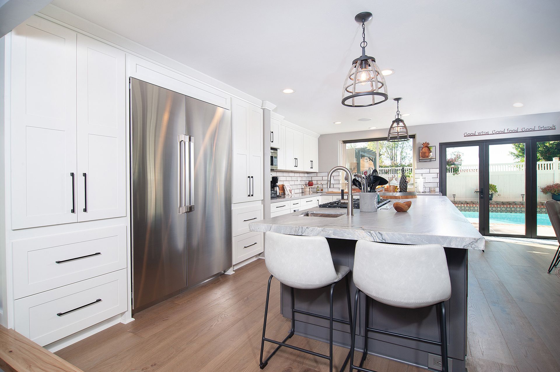 Modern white kitchen with large stainless steel fridge, island with seating, and view of a pool.