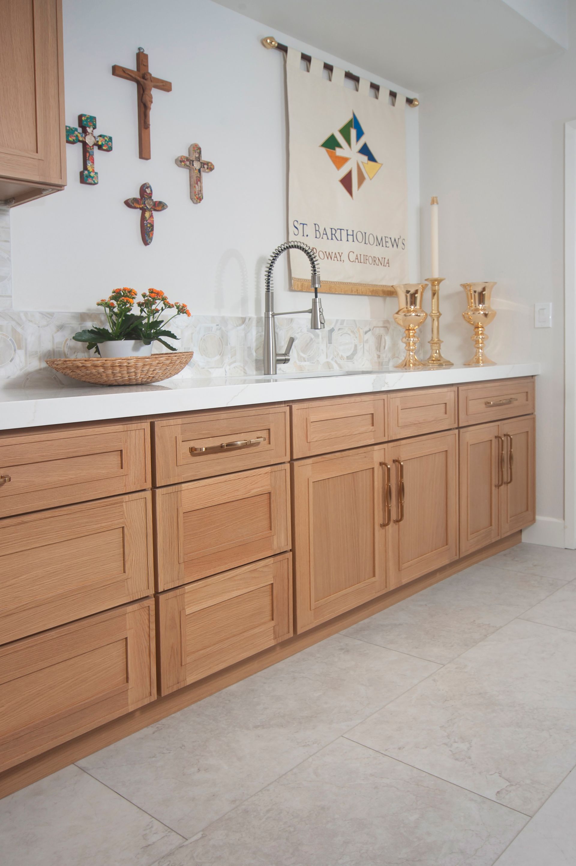 Kitchen with light wood cabinets, white countertops, religious crosses, banner, and sink.