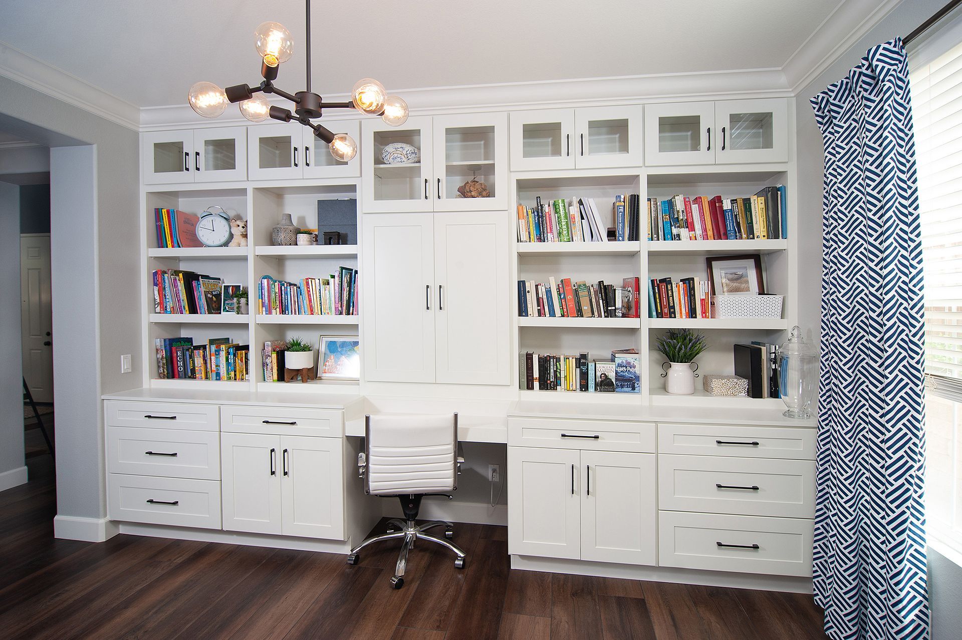 White built-in home office with bookshelves, desk, and cabinets against a gray wall, lit by an unusual chandelier.