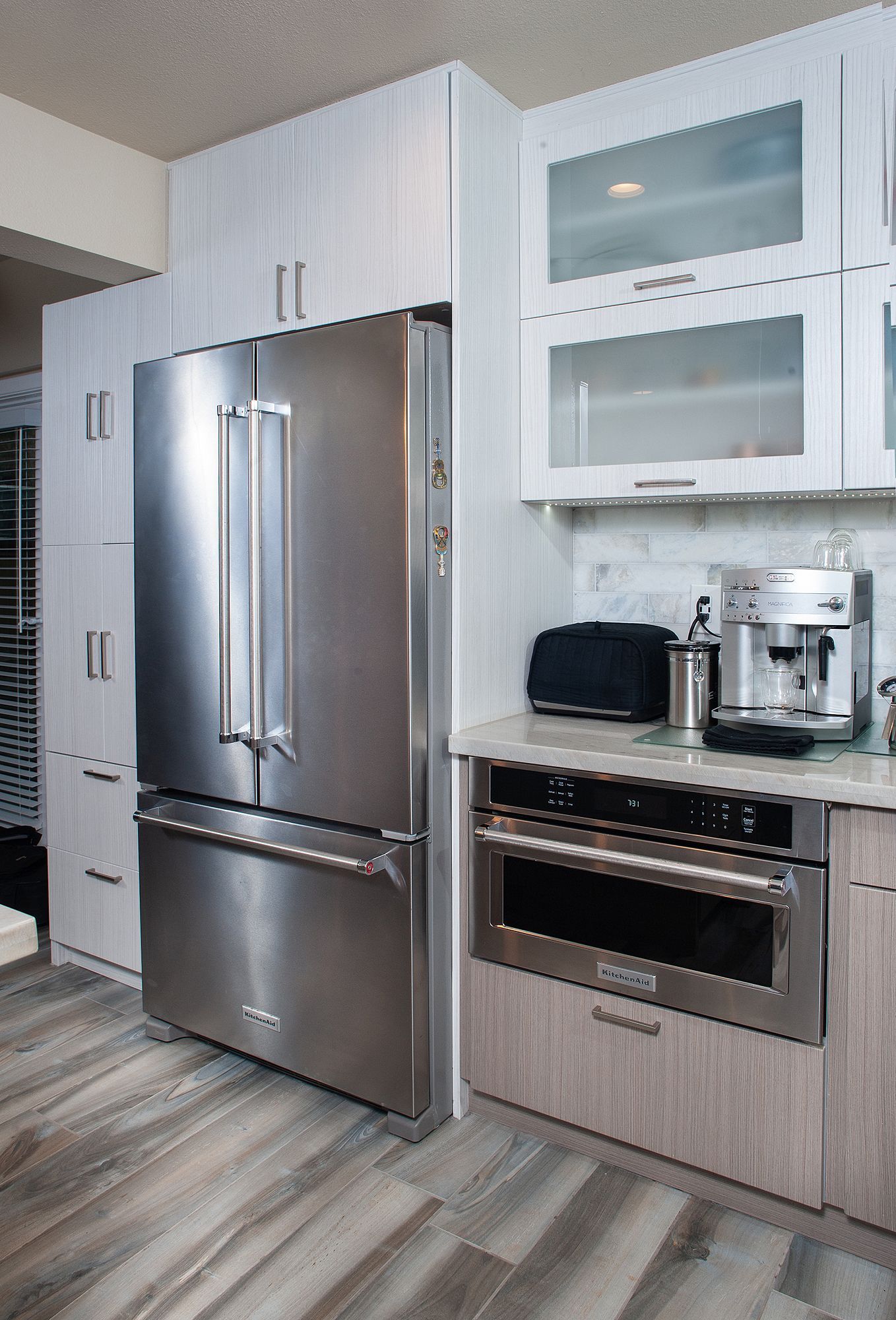 Stainless steel refrigerator and microwave built into a modern kitchen with light wood cabinets and flooring.