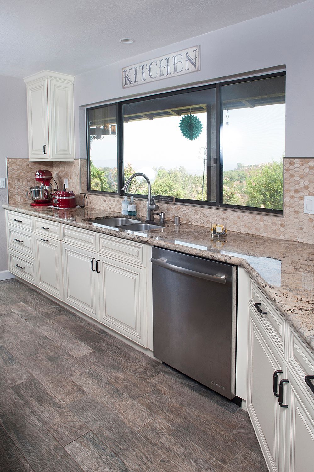 Cream-colored kitchen cabinets, stainless steel dishwasher, sink, and large window with a view, brown and tan countertops.