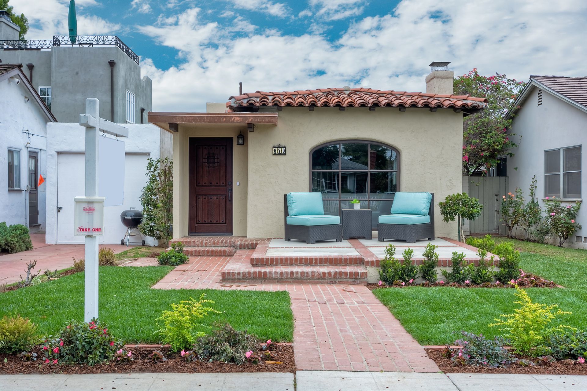 Small beige house with a red tile roof, front porch, and lawn. Blue cushions on outdoor seating.