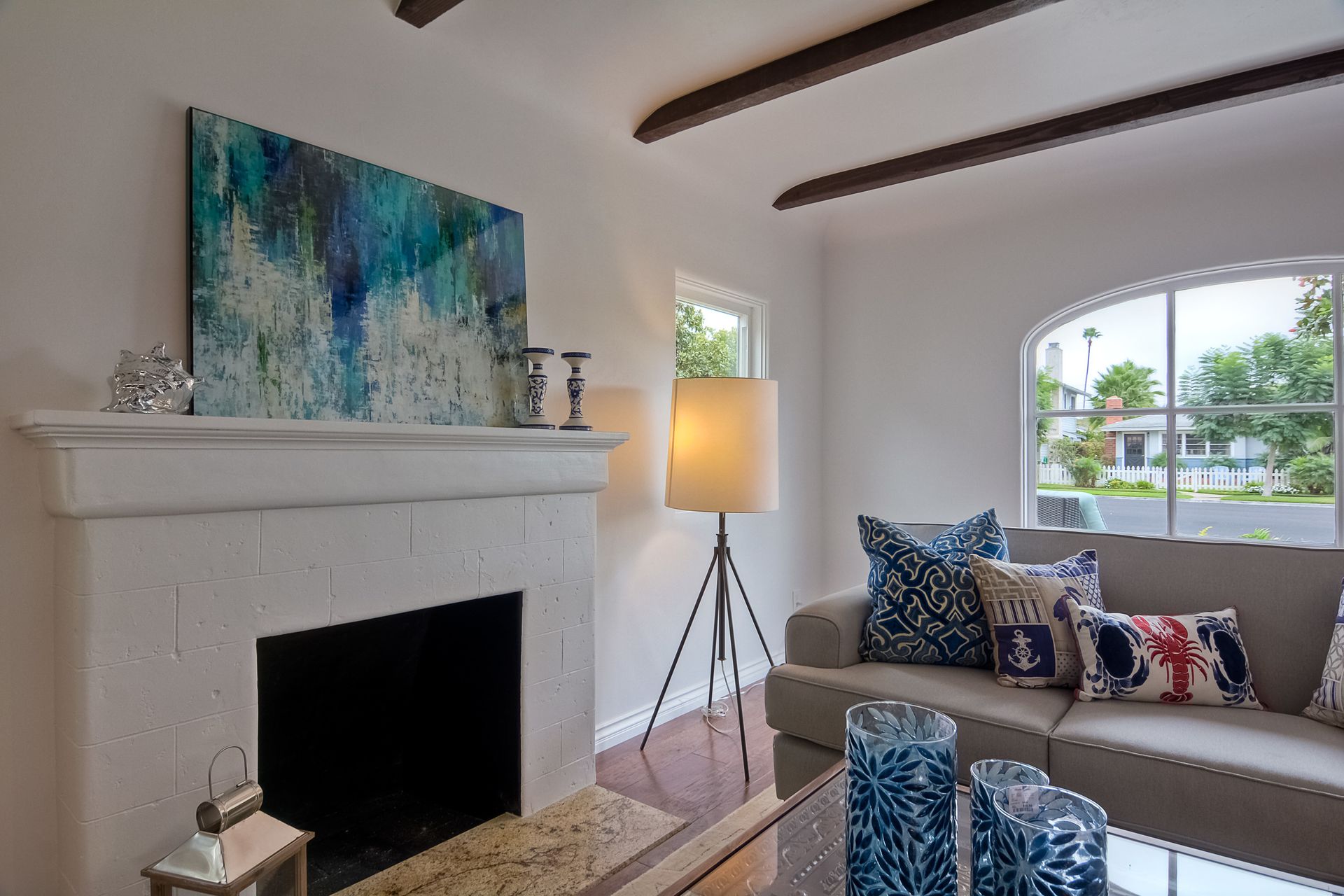 Living room with fireplace, sofa, and artwork. White walls, dark beams, colorful pillows, and floor lamp.