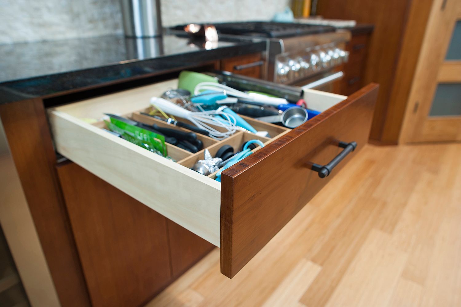 Kitchen drawer open, filled with utensils in wooden dividers. Brown cabinets and wood floor visible.