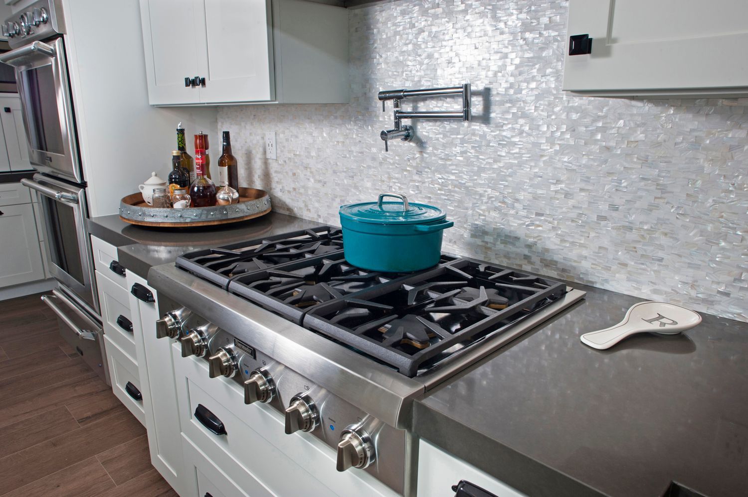 White kitchen with stainless steel stove and teal pot on a gas burner. Backsplash is iridescent tile.