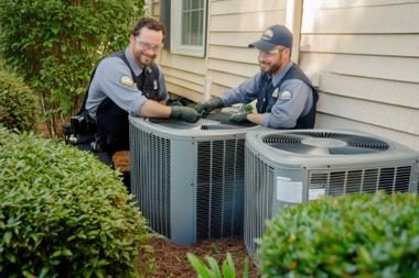 Two HVAC technicians inspecting outdoor air conditioning units beside a house and shrubs