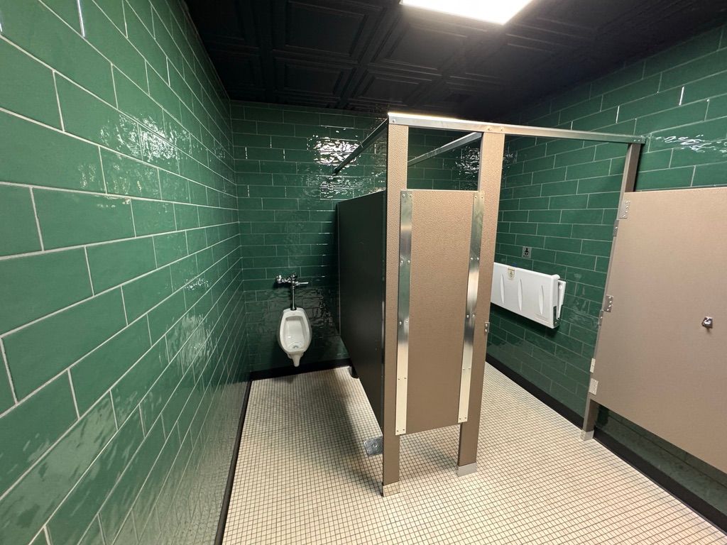 Public restroom stall area with green tiled walls, beige partitions, and a urinal visible in the back.