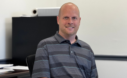Smiling man in a gray striped polo sitting in an office chair in a classroom or meeting room