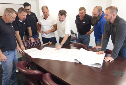 Group of men in a meeting room leaning over large blueprint plans on a conference table