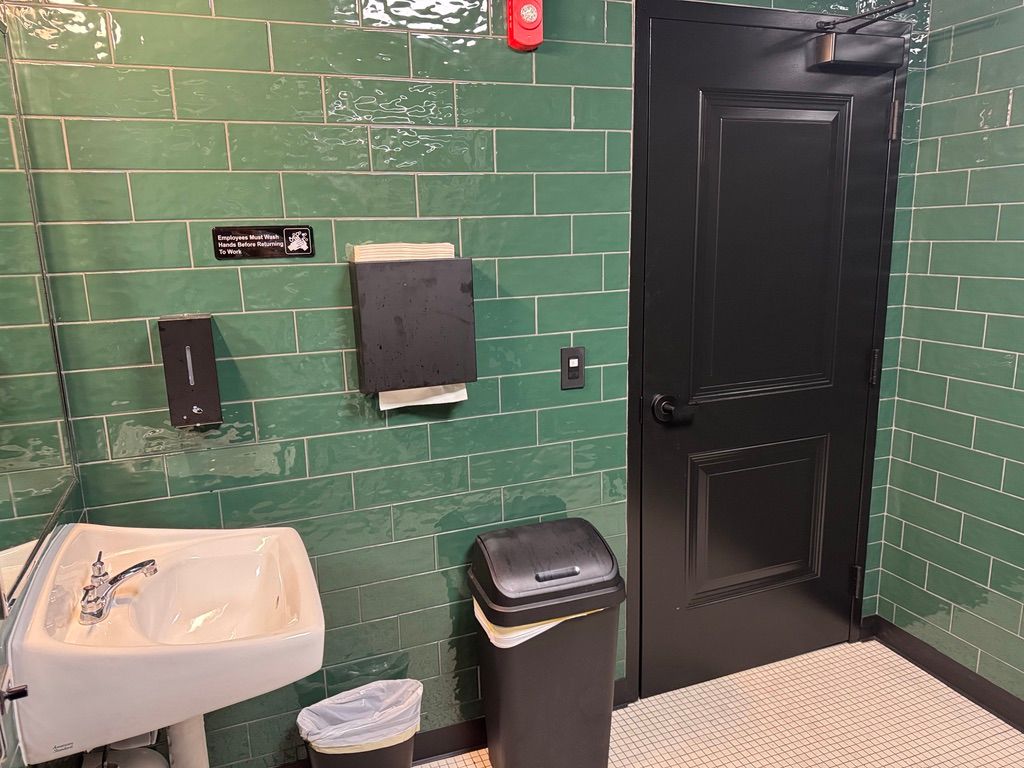 Bathroom with green tiled walls, black door, sink, paper towel dispenser, and trash can.