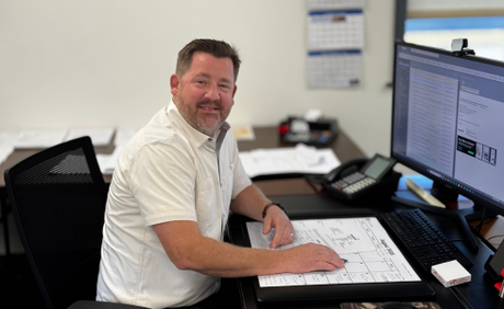 Man sitting at a desk with blueprints and dual monitors in a bright office, smiling at the camera