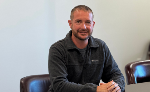 Man seated in a chair, smiling in a gray sweater against a plain wall.
