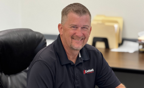 Smiling man in a black polo sitting at an office desk with a computer and files in the background