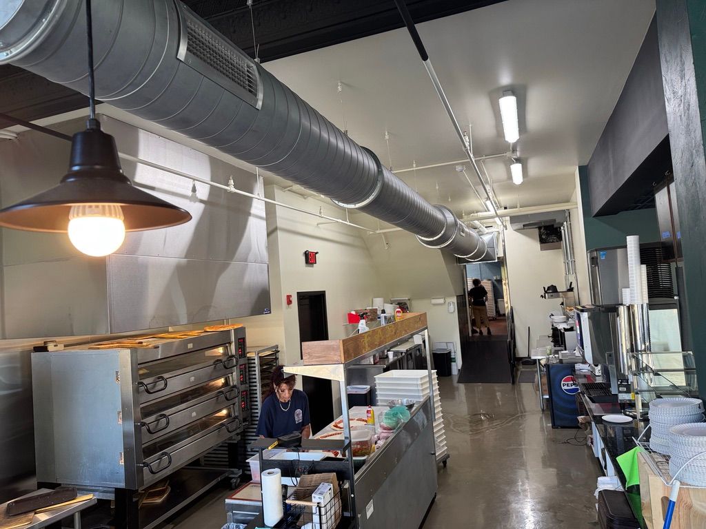 Commercial kitchen corridor with stainless steel counters, ovens, and overhead ductwork under bright lights