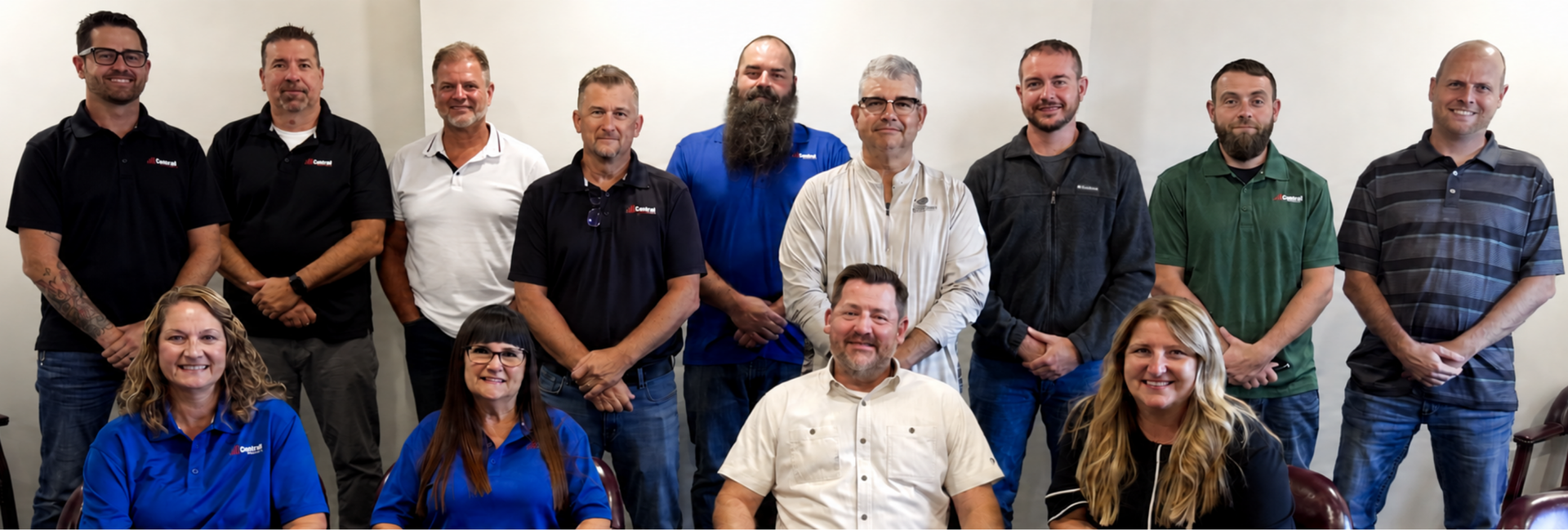 Group of coworkers posing indoors, standing and sitting in two rows against a plain wall.
