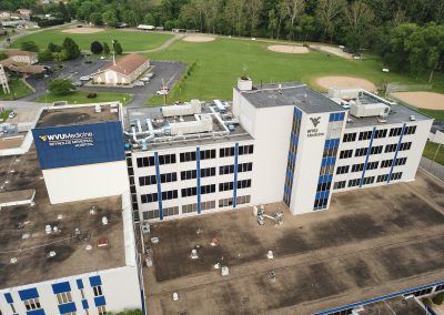 Aerial view of a multi-story white office building beside a green field and parking lot.