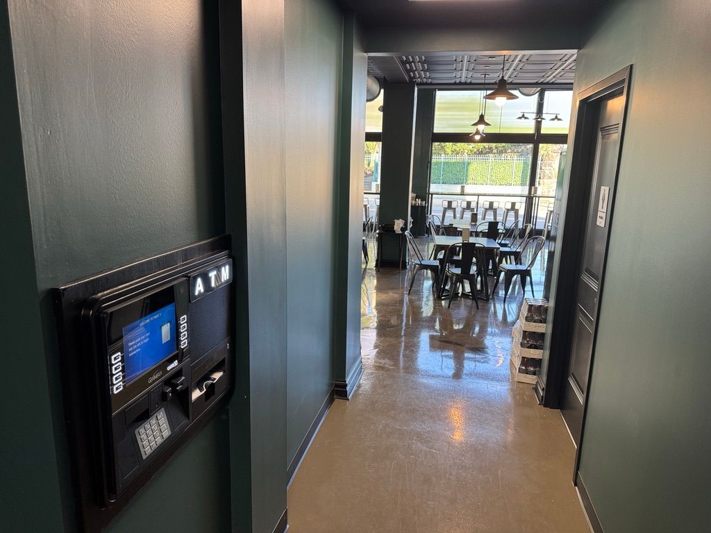 Hallway leading to a bright café with tables and chairs, viewed past a wall-mounted control panel.