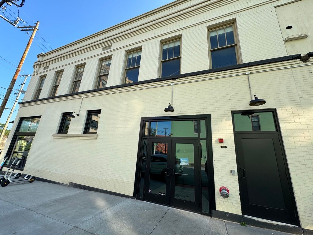 Cream brick storefront with black doors and windows on a sunny city sidewalk.
