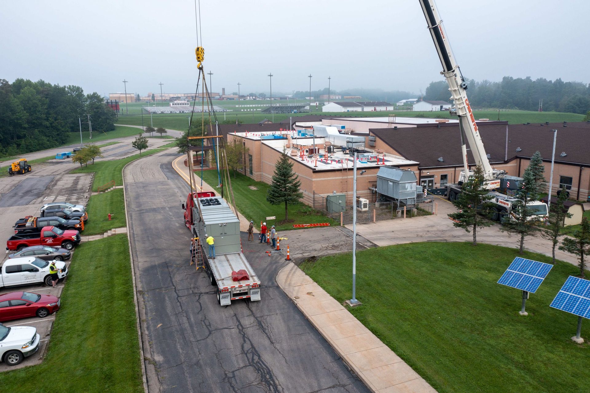 Construction workers and cranes beside a brown industrial building on a rainy roadside