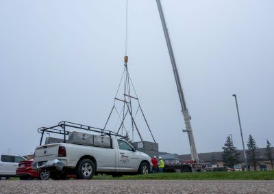 Crane lifting a white pickup truck with a roof rack in a parking lot on a cloudy day