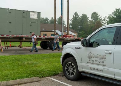 Workers walking beside a flatbed trailer and white pickup in a parking lot, with a large green container on the trailer