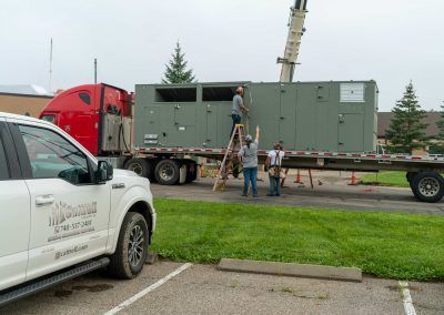 Workers using a ladder beside a large green utility trailer and red truck in a parking lot