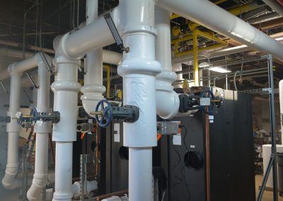Industrial pipes and valves in a mechanical room with white insulated piping and dark equipment underneath
