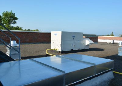 Rooftop HVAC unit beside a low brick wall, with shiny metal ductwork in the foreground.