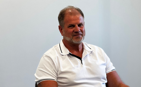 Man in a white polo shirt seated against a plain light-gray wall, looking slightly off-camera