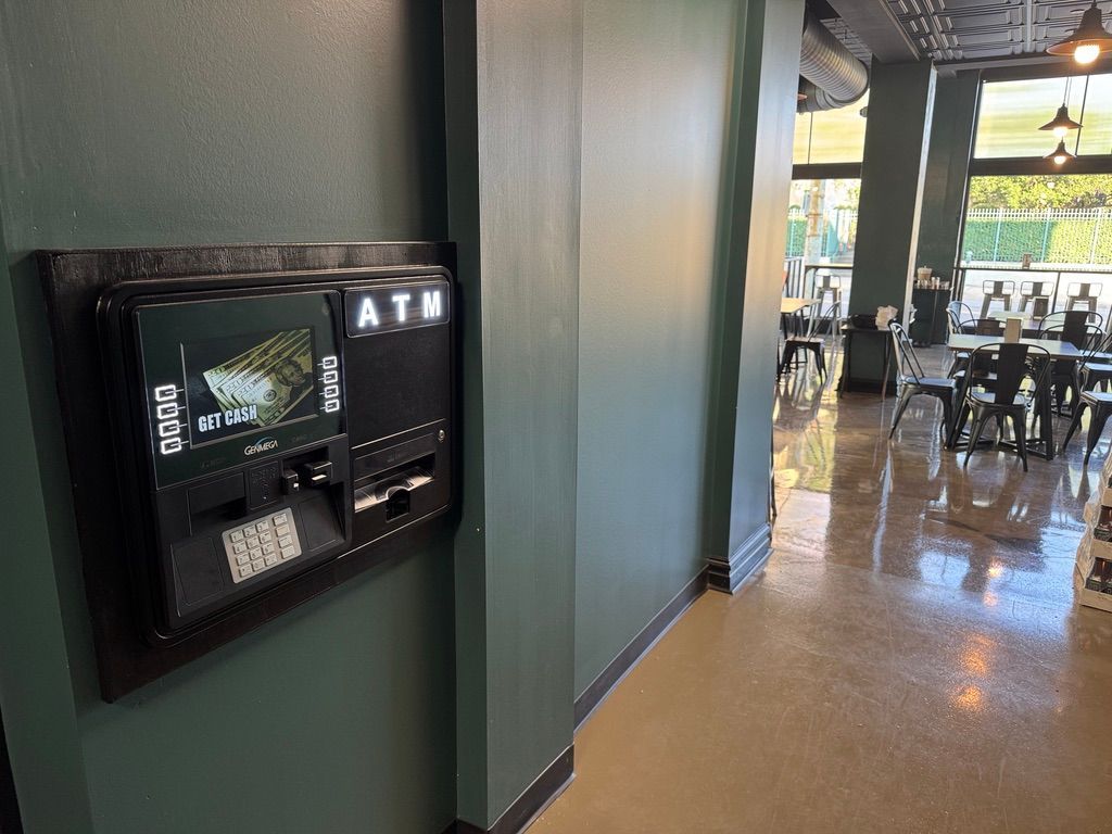 Wall-mounted black ATM on a green hallway wall, with a view into a bright restaurant beyond.