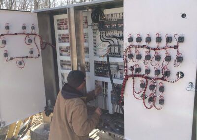 Technician working on an open electrical control panel with wires and gauges outdoors